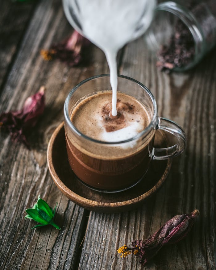 Glass cup of hot chocolate with milk pour and dried herbs on wooden table.